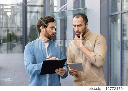 Two men are engaged in a discussion outdoors. One holds a clipboard, while the other considers a tablet in their hands. 127725704