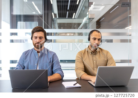 Two business professionals wearing headsets work diligently on their laptops in a modern office setting. They focus on their tasks intently. Two business professionals wearing headsets work diligently on their laptops in a modern office setting. They focus on their tasks intently. 127725720