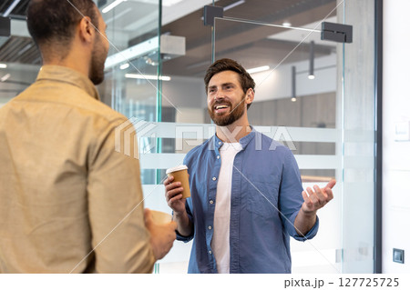 Two men are chatting in an office, one holding a coffee cup and gesturing while the other listens. Two men are chatting in an office, one holding a coffee cup and gesturing while the other listens. 127725725
