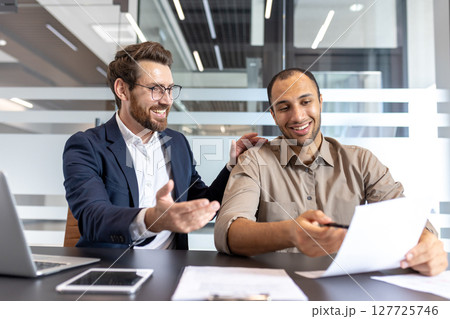 Two diverse businessmen smiling, reviewing paperwork, and discussing matters in a modern office environment. Collaboration and teamwork are evident. Two diverse businessmen smiling, reviewing paperwork, and discussing matters in a modern office environment. Collaboration and teamwork are evident. 127725746