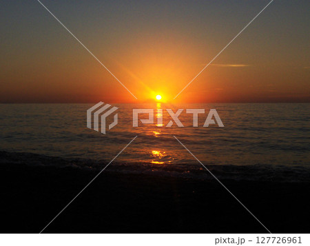 The breathtaking beauty of a sunset in Calabria, Italy. The sky is painted in rich hues of orange, pink, and gold as the sun dips below the horizon, casting a warm glow over the calm sea. 127726961