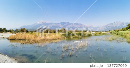 Serene landscape featuring calm river, lush greenery, and distant mountains under clear blue sky Serene landscape featuring calm river, lush greenery, and distant mountains under clear blue sky 127727069