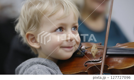 A young boy with blonde hair smiles while holding a violin, engaging in a music class at a community center. His concentration highlights the joy of learning an instrument. 127727096