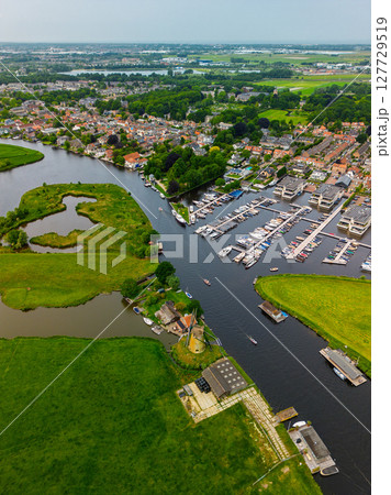 A top-down view of a marina with multiple boats docked along wooden piers, a boat cruising through a narrow waterway surrounded by green parks and natural grassy areas. A top-down view of a marina with multiple boats docked along wooden piers, a boat cruising through a narrow waterway surrounded by green parks and natural grassy areas. 127729519