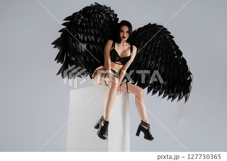 Woman in lingerie with striking black wings poses stylishly on a pedestal Woman in lingerie with striking black wings poses stylishly on a pedestal 127730365
