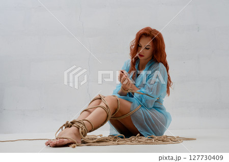 Red-haired woman elegantly bound in rope poses in a minimalist setting 127730409