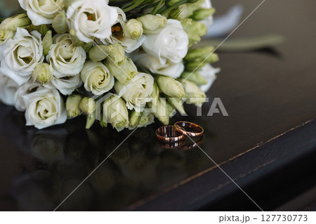 Wedding rings and white rose bouquet on dark wooden table in warm indoor lighting 127730773