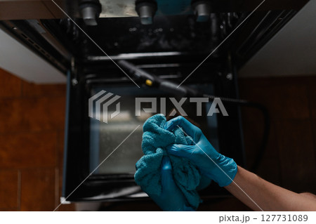 High-angle view of housekeeper male wearing blue gloves squeezing out wet rag after cleaning inside of gas oven with steam cleaner on blurred background. Concept of housekeeping and cleaning. High-angle view of housekeeper male wearing blue gloves squeezing out wet rag after cleaning inside of gas oven with steam cleaner on blurred background. Concept of housekeeping and cleaning. 127731089