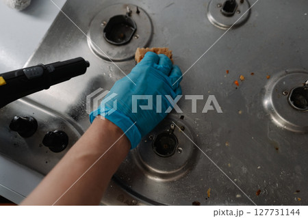 Top view of housekeeper wearing blue gloves using steam cleaner and sponge to remove grease and dirt from stainless steel gas stove, ensuring hygiene and cleanliness in kitchen, close-up. 127731144