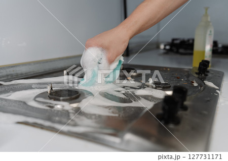 Male hand scrubbing gas stove with sponge and detergent, promoting hygiene and achieving sparkling clean kitchen surface tackling dirt and grease. Concept of domestic work, housekeeping and household. Male hand scrubbing gas stove with sponge and detergent, promoting hygiene and achieving sparkling clean kitchen surface tackling dirt and grease. Concept of domestic work, housekeeping and household. 127731171