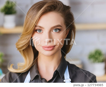 A woman with blonde hair and blue eyes wearing a gray shirt, standing indoors with a blurred background of shelves and plants 127734985