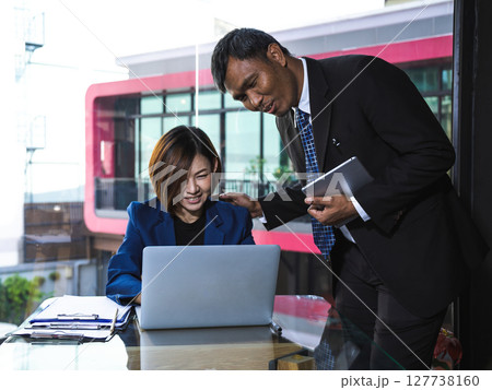 Businesswoman in navy suit smiling at laptop while businessman in black suit with tablet encourages and supports her by patting shoulder Businesswoman in navy suit smiling at laptop while businessman in black suit with tablet encourages and supports her by patting shoulder 127738160