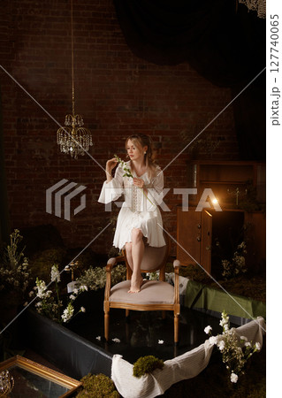 Woman in white dress sitting on chair in conceptual indoor swamp art setup 127740065