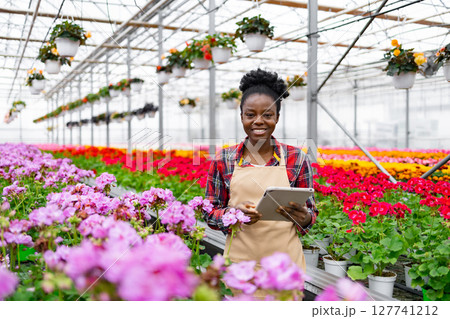 A smiling African woman in a greenhouse with a tablet, surrounded by vibrant flowers and plants, and other potted plants. 127741212