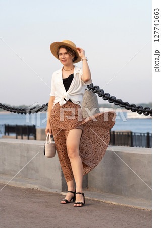 Cheerful woman holding straw hat enjoys breezy waterfront stroll. Cheerful woman holding straw hat enjoys breezy waterfront stroll. 127741663