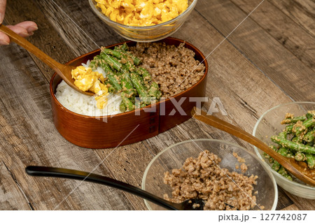 三食丼弁当を詰める そぼろ,いんげんの胡麻和え, 炒り卵をご飯に乗せる 三食丼弁当を詰める そぼろ,いんげんの胡麻和え, 炒り卵をご飯に乗せる 127742087