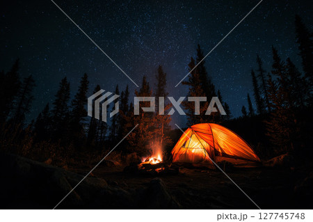 Cozy tent glowing warmly under a starry night sky in a tranquil forest setting. 127745748