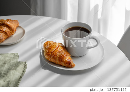 Cup of espresso coffee and fresh croissant on a plate on white table near window with shadows. 127751135