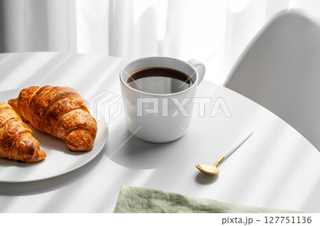 Cup of espresso coffee and fresh croissants on a plate on white table near window with shadows 127751136