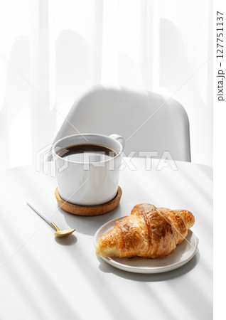 Cup of espresso coffee and fresh croissant on white table near window with morning shadows 127751137