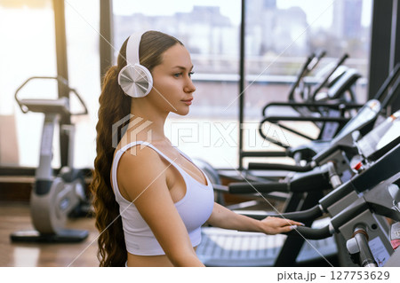Young sporty woman running on machine in the gym and listening music in headphones Young sporty woman running on machine in the gym and listening music in headphones 127753629