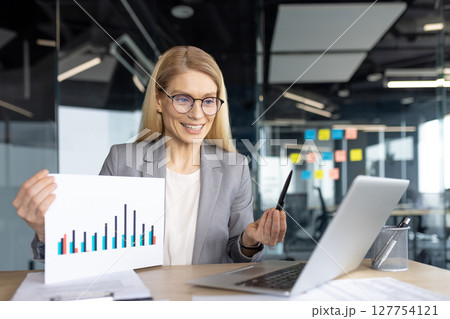 Professional woman presenting a bar chart via video conference from her office, using a laptop. She is wearing a grey blazer and glasses. 127754121