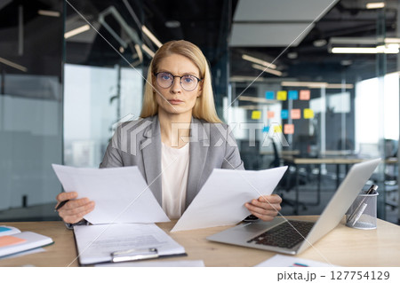 A businesswoman in a professional setting sits at her desk, reviewing documents with a serious expression. She is wearing glasses. 127754129