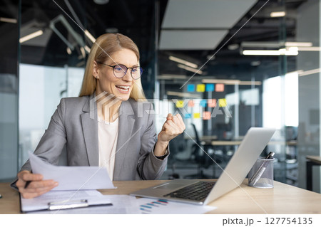 A businesswoman celebrates her success, holding documents and viewing a laptop screen. The office environment suggests a work-related achievement or positive outcome. A businesswoman celebrates her success, holding documents and viewing a laptop screen. The office environment suggests a work-related achievement or positive outcome. 127754135