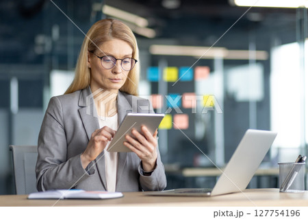 A businesswoman in glasses reviews a tablet at her desk, working in a modern office setting. 127754196