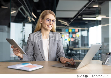 A businesswoman works in an office setting, utilizing a laptop and tablet simultaneously. She smiles while appearing to be enjoying her workday. 127754202