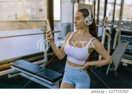 Portrait of young sportswoman with smartphone listening to music in gym. Portrait of young sportswoman with smartphone listening to music in gym. 127755767