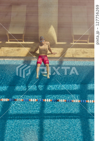 Man relaxing at edge of indoor swimming pool in red shorts with sunlit reflections on water 127756269