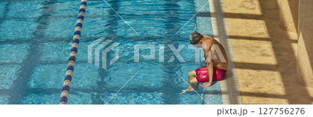 Man in swimwear sitting on pool edge dipping toes in clear blue water 127756276