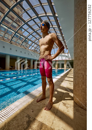 Competitive swimmer in bright swimwear stands confidently beside indoor pool under arched glass Competitive swimmer in bright swimwear stands confidently beside indoor pool under arched glass 127756282