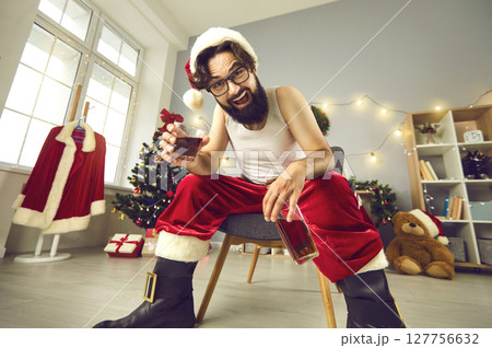 Happy man in undershirt and Santa costume drinking whiskey at Christmas party at home 127756632