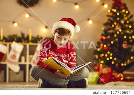 Positive boy in Santa hat and red traditional sweater sitting on floor and reading book Positive boy in Santa hat and red traditional sweater sitting on floor and reading book 127756694