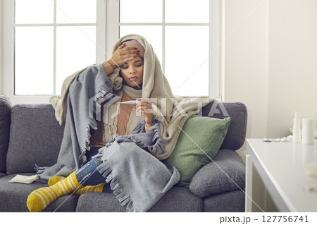 Sick young woman, wrapped in blankets, looking at thermometer after taking her temperature 127756741