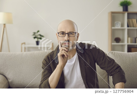 Webcam view of serious adult man sitting on sofa at home, looking at camera and thinking 127756904
