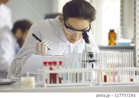 Woman in a white coat looks in a microscope. 127757293