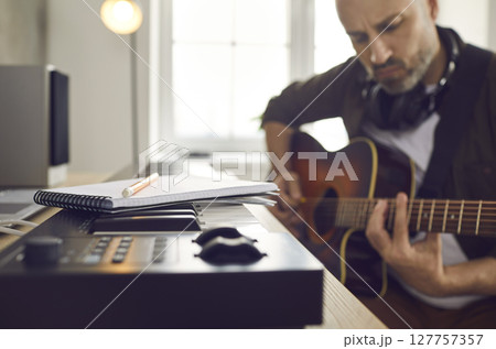 Musician playing guitar and writing music while sitting by keyboard with paper and pencil 127757357