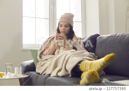 Sick young woman suffering from bad cold or flu, sitting wrapped in blanket, and drinking hot tea 127757380