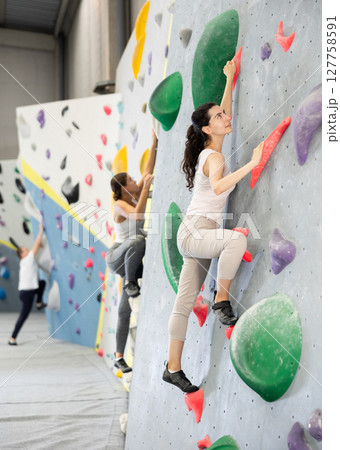 Girl is mastering climbing on training wall in gym. Woman holds on tightly to ledges and strives for top of bouldering route Girl is mastering climbing on training wall in gym. Woman holds on tightly to ledges and strives for top of bouldering route 127758591