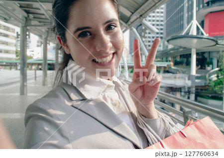 A cheerful woman in a suit enjoys urban life, holding shopping bags and flashing a peace sign outdoors. A cheerful woman in a suit enjoys urban life, holding shopping bags and flashing a peace sign outdoors. 127760634