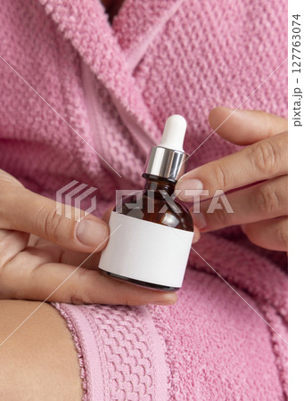 Woman in pink bathrobe with glass dropper bottle with label in hands closeup, cosmetic mockup Woman in pink bathrobe with glass dropper bottle with label in hands closeup, cosmetic mockup 127763074