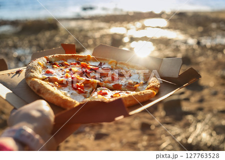 Pizza with tomatoes and mozzarella lies in brown paper box in female hand on beach sun day 127763528