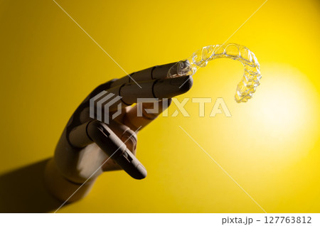 Wooden hand of an impersonal mannequin holds transparent polymer dental aligners on a colored background for teeth alignment. Aesthetic and comfortable teeth alignment Wooden hand of an impersonal mannequin holds transparent polymer dental aligners on a colored background for teeth alignment. Aesthetic and comfortable teeth alignment 127763812