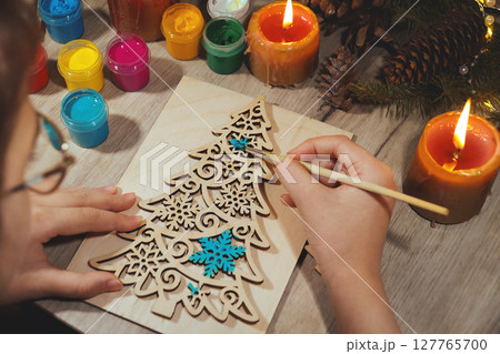 Children's hands paint wooden toy Christmas tree with spruce branches, pinecones and candles on table 127765700