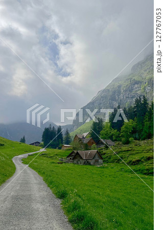Axalp region of Bern canton Swiss rural landscape winds through lush green grass, guiding toward cozy cabins nestled against majestic mountains. 127767053
