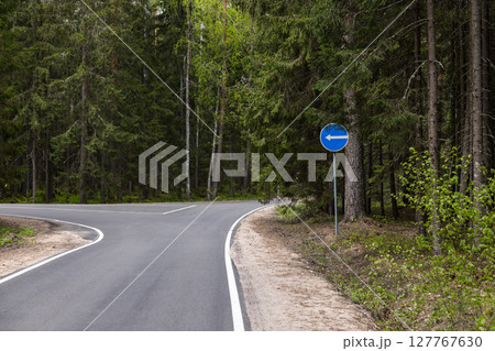 An empty asphalt road winding through a green forest, traffic sign directing left An empty asphalt road winding through a green forest, traffic sign directing left 127767630