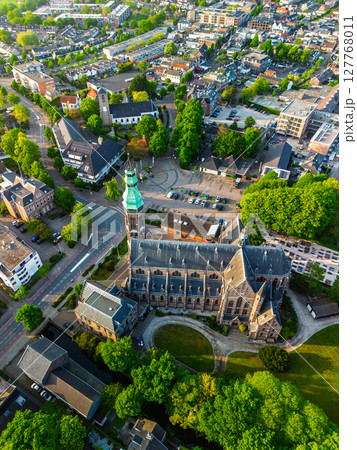 Aerial view of a picturesque European town featuring a prominent church with a tall green spire surrounded by lush greenery, quaint houses, and a town square with parked cars. Aerial view of a picturesque European town featuring a prominent church with a tall green spire surrounded by lush greenery, quaint houses, and a town square with parked cars. 127768011